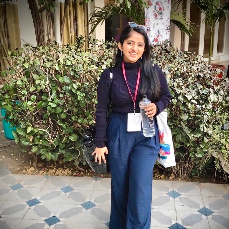prakruti maniar profile photo and personal website - girl in blue pants and blue sweater, holding a botle standing with a smile against shrubs. hair is left open, specs are being used as a hairband. the head has a left tilt